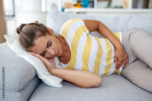Woman lying on sofa looking sick in the living room. Beautiful young woman lying on bed and holding hands on her stomach. Woman having painful stomachache on bed, Menstrual period photo