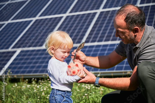 Modern father and his little son saving money in piggy bank. Adorable kid learning how to save money. Man and his cute child putting some cash to piggy bank on background of solar panels. © anatoliy_gleb