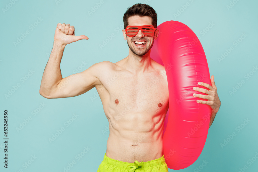 Young sexy man wear green shorts swimsuit relax near hotel pool hold inflatable rubber ring point thumb finger on himself isolated on plain blue background. Summer vacation sea rest sun tan concept.