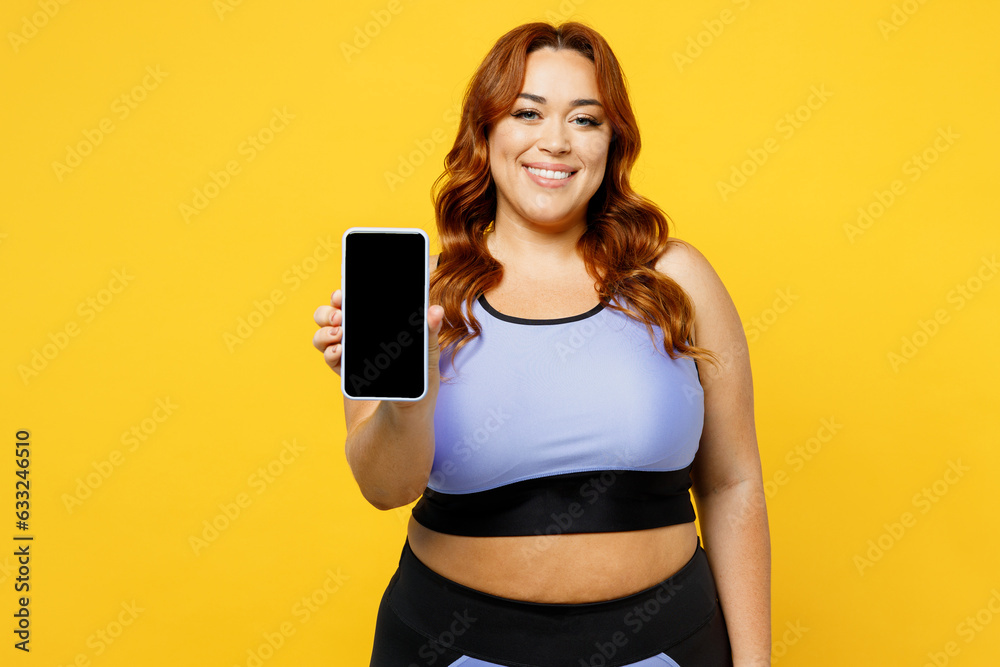 Young chubby overweight plus size big fat fit woman wear blue top warm up training hold use blank screen mobile cell phone isolated on plain yellow background studio home gym. Workout sport concept. Young chubby overweight plus size big fat fit woman wear blue top warm up training hold use blank screen mobile cell phone isolated on plain yellow background studio home gym. Workout sport concept.