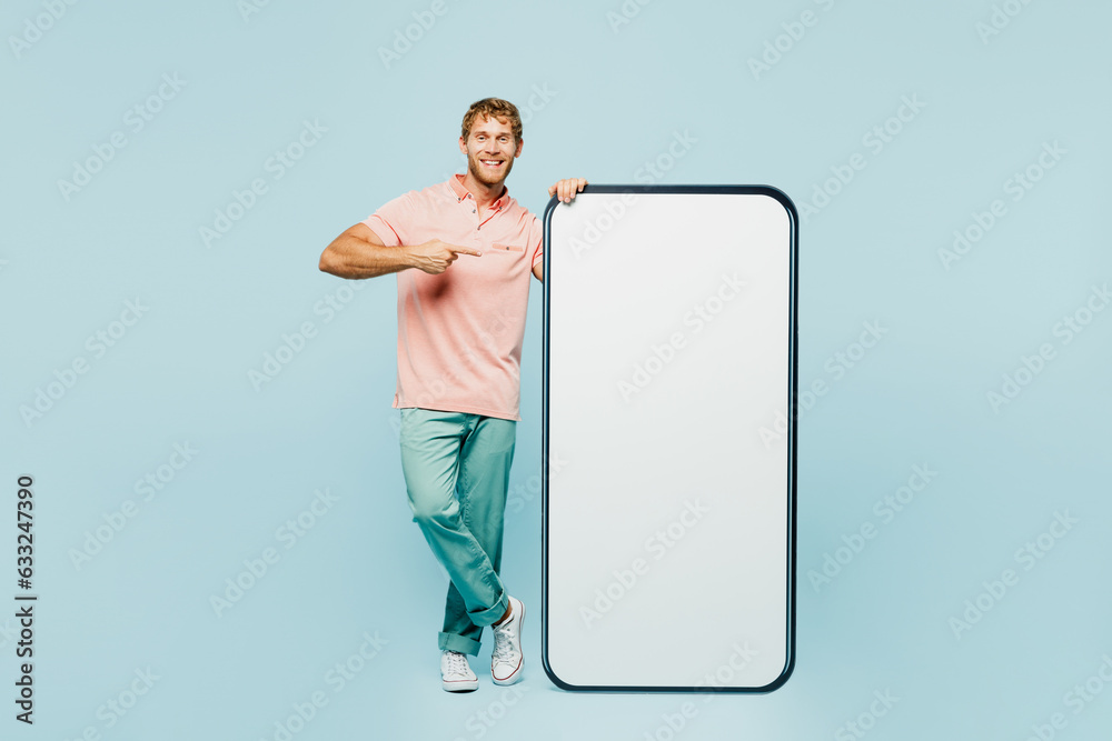 Full body young man wear pink t-shirt casual clothes point index finger on big huge blank screen mobile cell phone smartphone with workspace area isolated on plain blue background. Lifestyle concept. Full body young man wear pink t-shirt casual clothes point index finger on big huge blank screen mobile cell phone smartphone with workspace area isolated on plain blue background. Lifestyle concept.