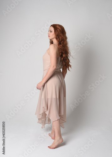 Full length portrait of beautiful female model with long brunette hair wearing a creamy pink gown dress. graceful dancing pose, with gestural hands isolated on white studio background. 