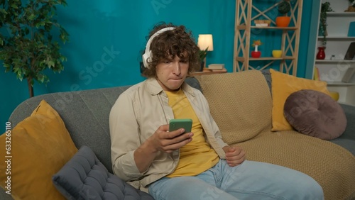 Young man in headphones sitting on the couch in the living room and listening to music on smartphone