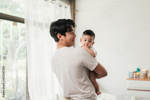 Happiness of fatherhood. Young Asian dad with adorable baby on his hands standing near window at home. Loving father spending time with infant child. Fathers day concept.