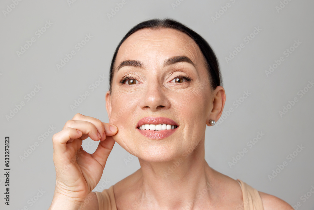 Close up beautiful mature woman pulling perfect skin and smiling a camera over grey background.