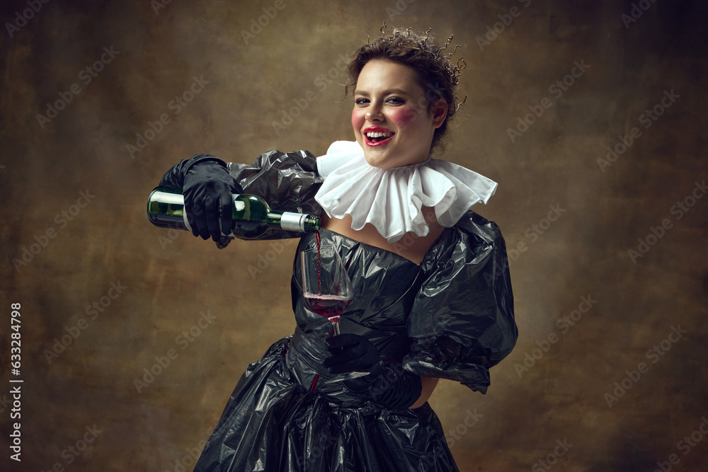 Portrait of cheerful woman in historic dress made of garbage bags, holding wine glass in hand and pour wine into it over vintage background.