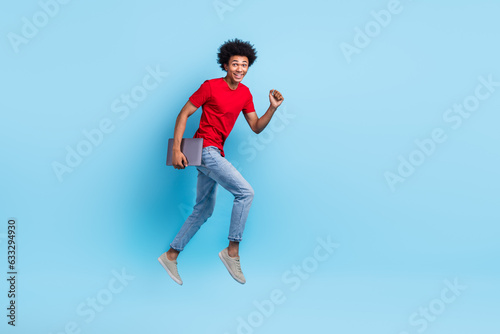 Full size photo of handsome guy dressed red stylish t-shirt denim pants hold laptop run to empty space isolated on blue color background