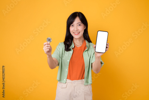 the innovation of future finance with young Asian woman in her 30s, elegantly dressed in orange shirt and green jumper, using smartphone screen display and crypto currency coin on yellow background.