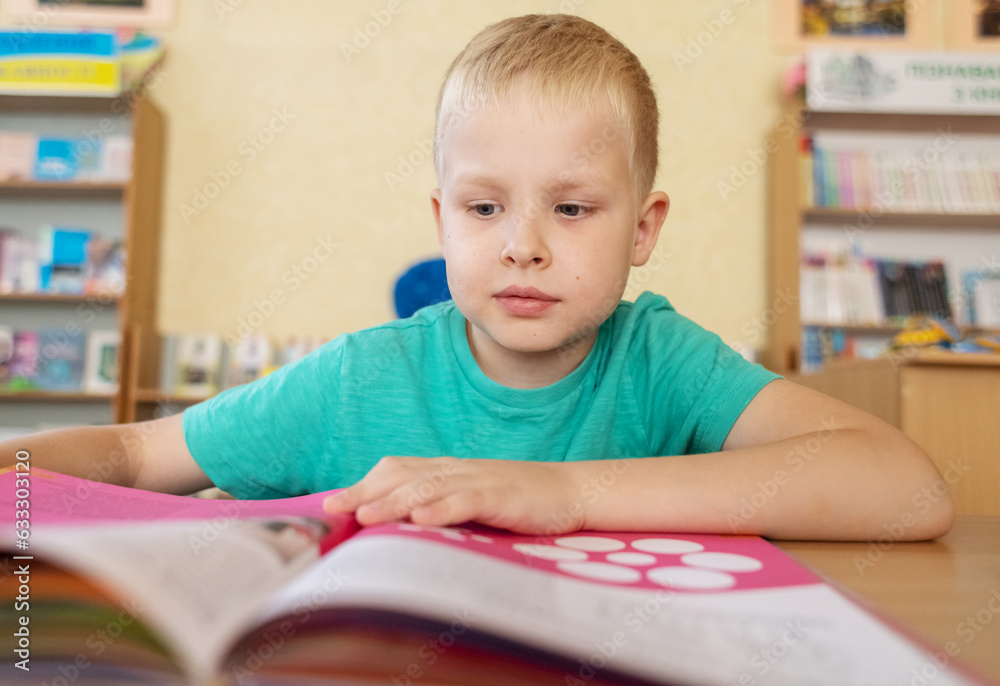 Boy in library. Seven year Ukrainian guy is sitting at a table in library and reading book. Concept of normal learning, thirst for knowledge, desire to learn new things. Blurred background and letters