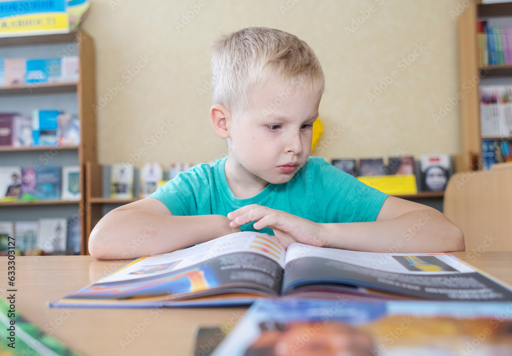 Boy in library. Seven year Ukrainian guy is sitting at a table in library and reading book. Concept of normal learning, thirst for knowledge, desire to learn new things. Blurred background and letters Boy in library. Seven year Ukrainian guy is sitting at a table in library and reading book. Concept of normal learning, thirst for knowledge, desire to learn new things. Blurred background and letters