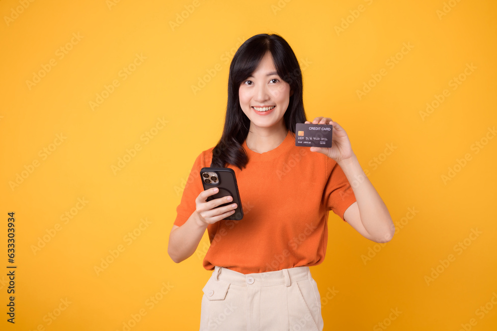 Enthusiastic young Asian woman in her 30s, using smartphone and holding credit card on yellow background. Swift online shopping. Enthusiastic young Asian woman in her 30s, using smartphone and holding credit card on yellow background. Swift online shopping.