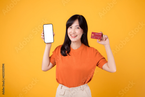 Vibrant Asian woman in her 30s, showcasing smartphone and credit card on yellow background. Effortless online payment shopping.