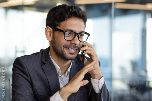 Young successful arab businessman boss talking on phone close up, man smiling contentedly inside office at workplace in business suit
