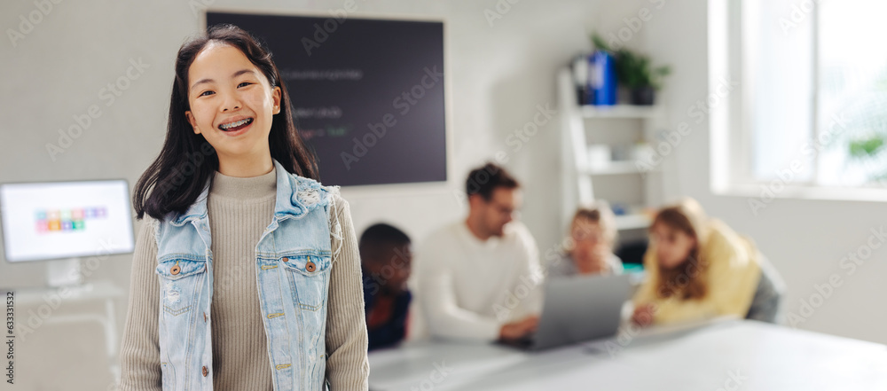 Fototapeta premium Young Asian student smiling at the camera in a coding class, with her teacher and classmates in the background