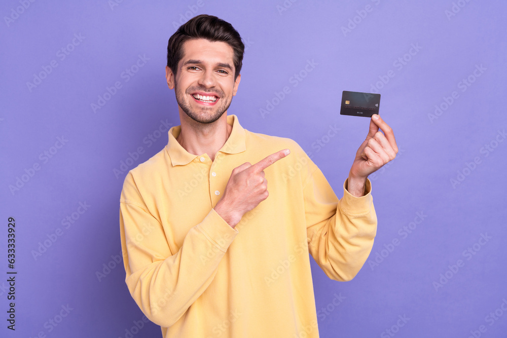 Photo of positive attractive guy beaming smile indicate finger arm hold debit card isolated on violet color background Photo of positive attractive guy beaming smile indicate finger arm hold debit card isolated on violet color background