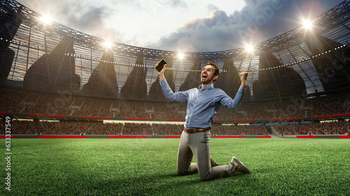 Leinwand Poster Man standing on knees on football arena, holding mobile phone and celebrating win of money