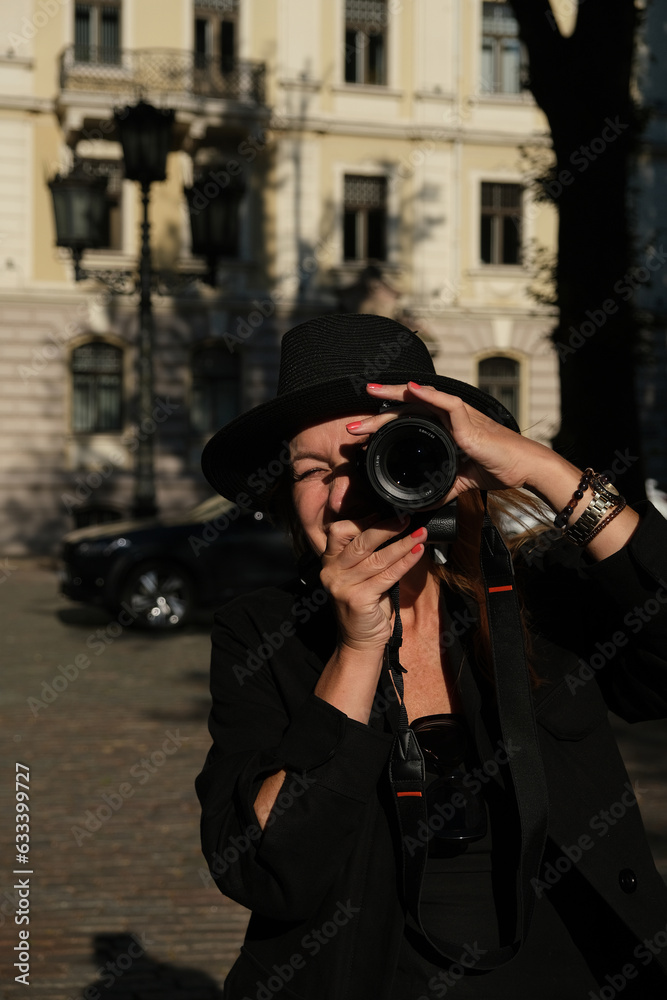 A woman takes pictures with a professional photo camera. A woman dressed in black clothes. Black wide brim. The woman is smiling. City in the background.
