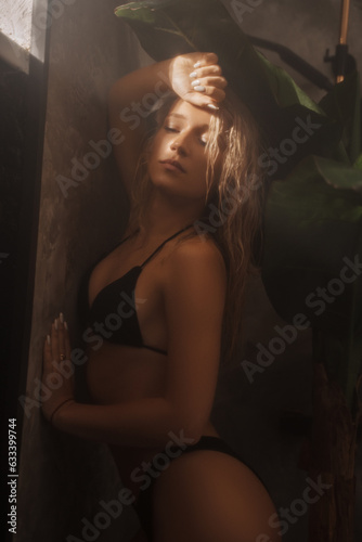Stunning model in a black swimsuit striking a pose in a shower surrounded by palm leaves, tropical concept, her face illuminated by bright sunlight