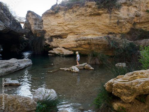 a little boy is climbing on a rock in the middle of the river.  Usero waterfall in Bullas, Murcia, Spain. He is wearing summer clothes and a hat to protect himself from the sun.