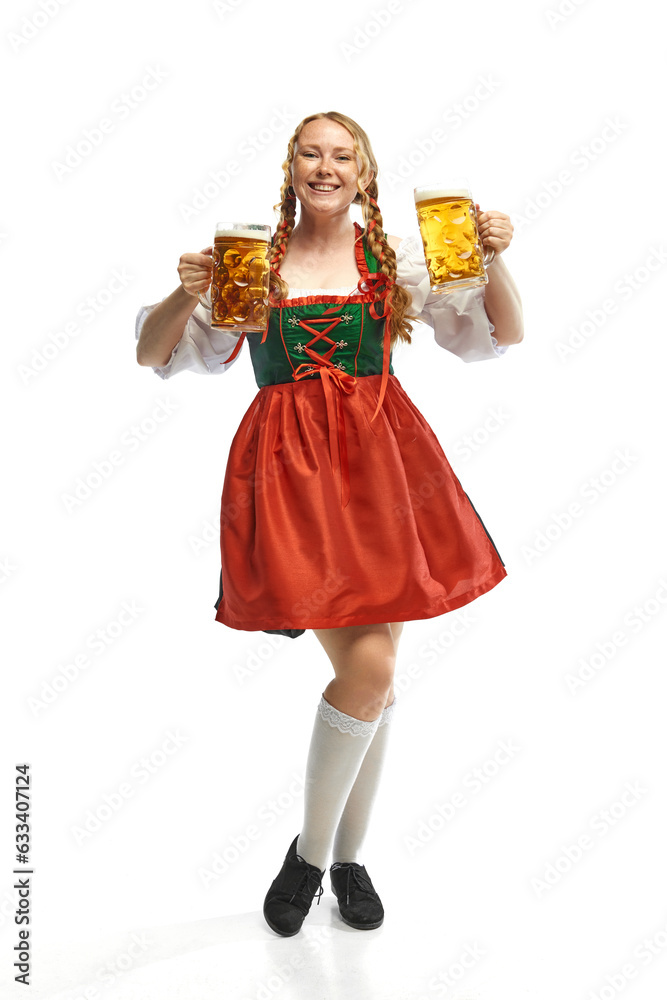 Full-lenght portrait of friendly young woman wearing folk German dirndl with two beer mugs over white background. Holiday event, Oktoberfest