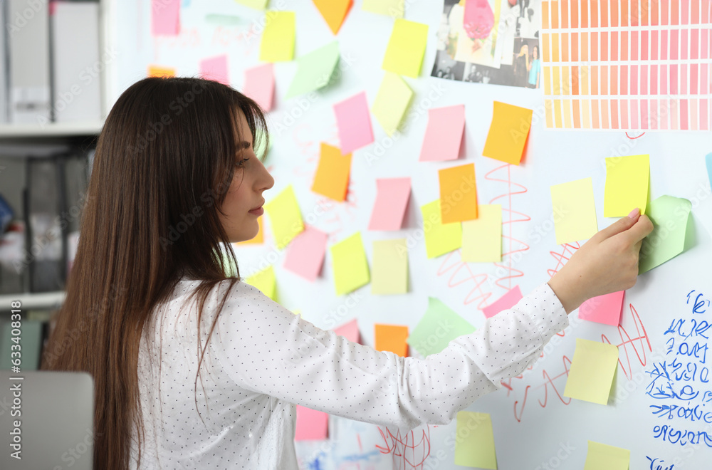 Portrait of pretty designer standing at workplace and making notes with colorful empty stickers. Beautiful businesswoman working on art project. Blurred background Portrait of pretty designer standing at workplace and making notes with colorful empty stickers. Beautiful businesswoman working on art project. Blurred background