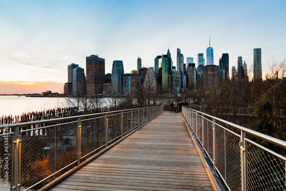 View of the skyscrapers of Lower Manhattan from a walkway path bridge in Brooklyn, lit by sunset.