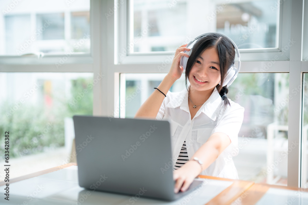 Happy of cute smiles Asian of attractive young Cute girl little wearing headphones and listening music using laptop computer working from at the cafe.Online education, elearning concept. Happy of cute smiles Asian of attractive young Cute girl little wearing headphones and listening music using laptop computer working from at the cafe.Online education, elearning concept.