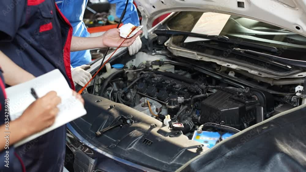 Selective focus, cropped view of a young male mechanic in uniform, standing in front of the car with an open hood, pulling out the dipstick and wipe off the oil with a cloth for checking the oil level Selective focus, cropped view of a young male mechanic in uniform, standing in front of the car with an open hood, pulling out the dipstick and wipe off the oil with a cloth for checking the oil level