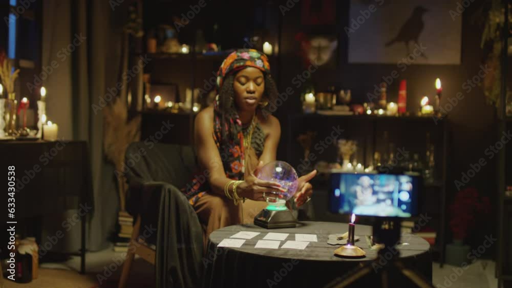 Medium long shot of African American seer videoblogging session with crystal ball when sitting at table in her studio in dark colors Medium long shot of African American seer videoblogging session with crystal ball when sitting at table in her studio in dark colors