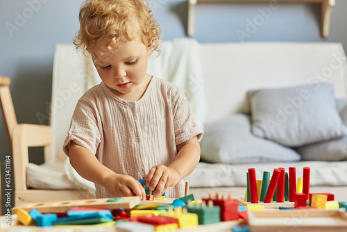 Kindergarten room activities. Colorful educational games. Small child's big curiosity. Concentrated wavy haired blonde toddler baby playing wooden educational toys at home interior.