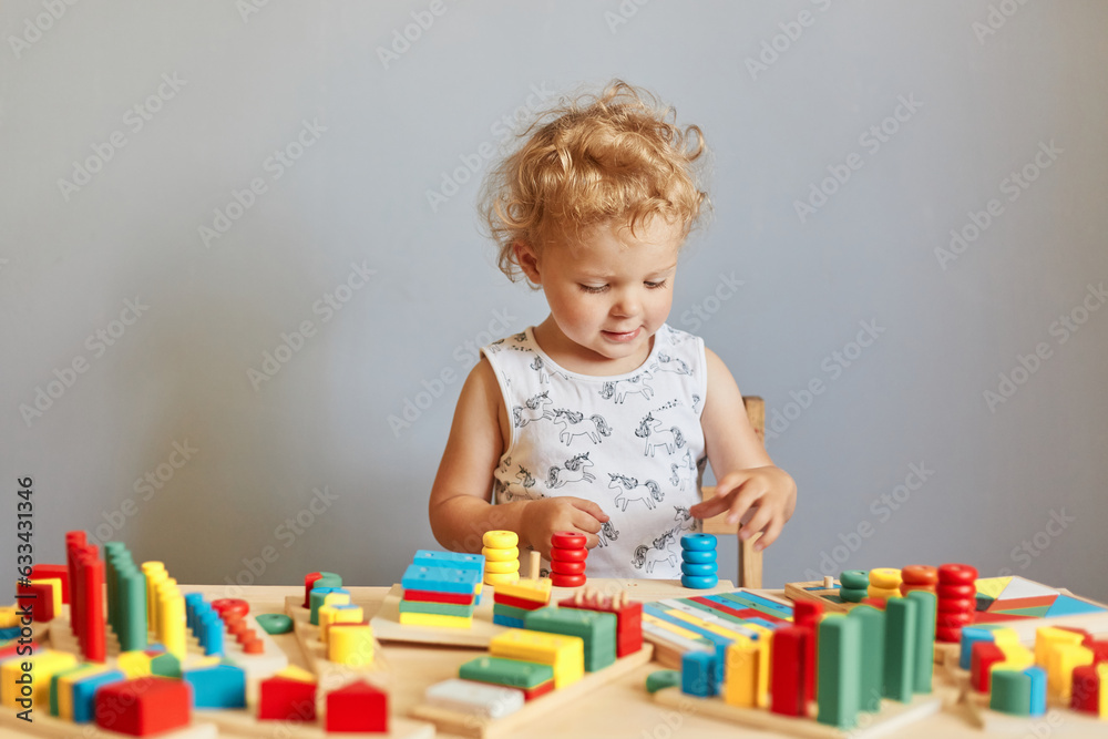 Curious child's playful learning. Kindergarten room activities. Building a playful preschooler. Cute wavy haired blonde toddler baby playing wooden educational toys isolated over gray background Curious child's playful learning. Kindergarten room activities. Building a playful preschooler. Cute wavy haired blonde toddler baby playing wooden educational toys isolated over gray background
