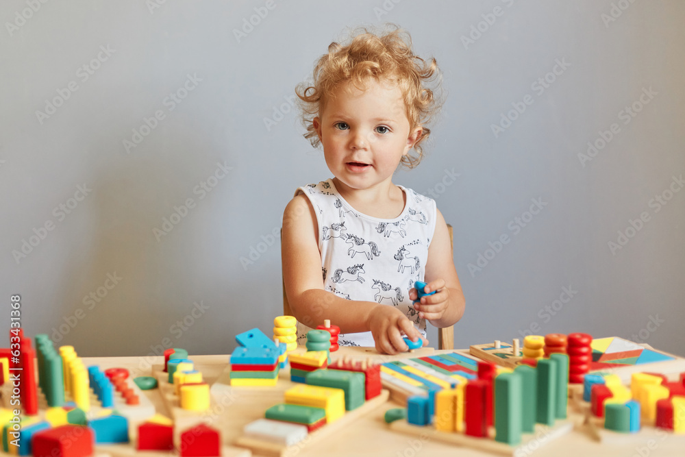Infant's daycare adventures. Learning through colorful play. Little nursery exploration. Funny wavy haired blonde toddler baby playing wooden educational toys isolated over gray background Infant's daycare adventures. Learning through colorful play. Little nursery exploration. Funny wavy haired blonde toddler baby playing wooden educational toys isolated over gray background