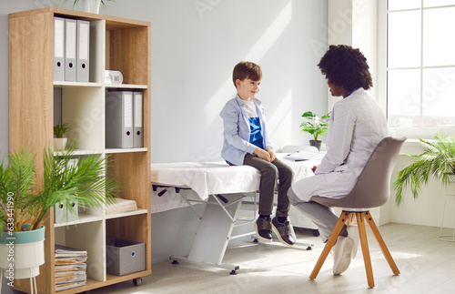 Female african american doctor talking with child patient on the couch during medical exam and listening complaints in clinic. Pediatrician checking a young boy and giving consultation in exam room.