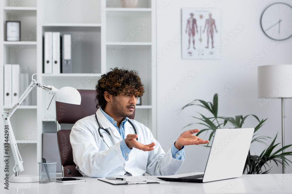 Young indian displeased and serious doctor inside office workplace talking remotely consulting patients using laptop for remote meeting