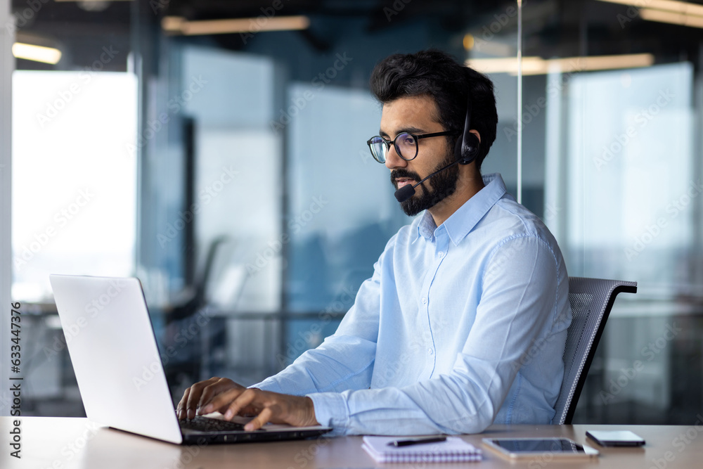 A young Indian businessman is concentrating on a laptop at an office table, wearing a headset, talking, negotiating, consulting A young Indian businessman is concentrating on a laptop at an office table, wearing a headset, talking, negotiating, consulting