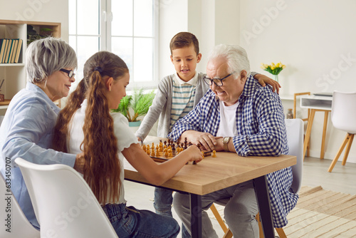 Grandparents and grandchildren spend time together, play chess and have fun. Happy old, retired granddad, grandma and little children sitting at table and playing game of chess. Family leisure concept photo