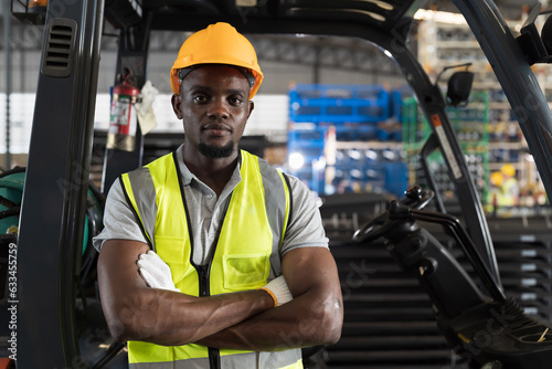 Portrait of African American male warehouse worker in safety vest and helmet standing after driving and operating on forklift truck for transfer products or parcel goods in the industrial storage ware