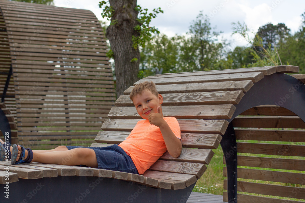 Child boy enjoys summer lying on a wooden sun lounger on the lake and shows a thumbs up. On a sunny day, a child is relaxing on a wooden deck chair by the lake.