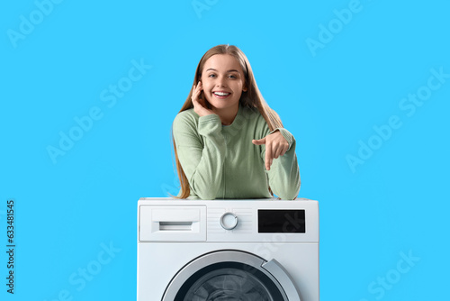 Pretty young woman leaning on washing machine and pointing at it on blue background