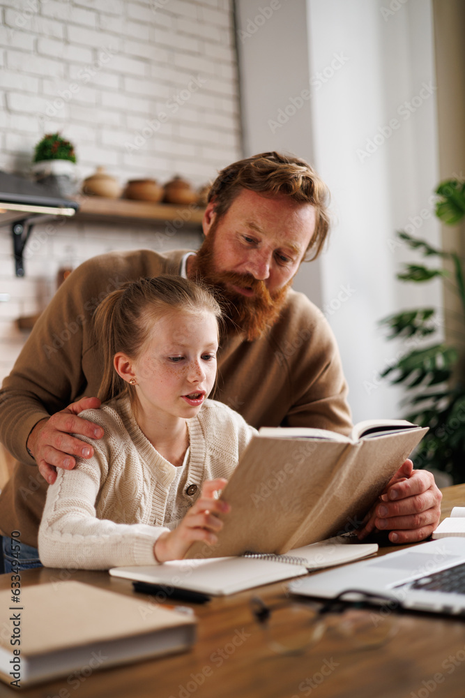 Fototapeta premium Bearded father hugging daughter reading book near laptop and notebook during e-learning at home