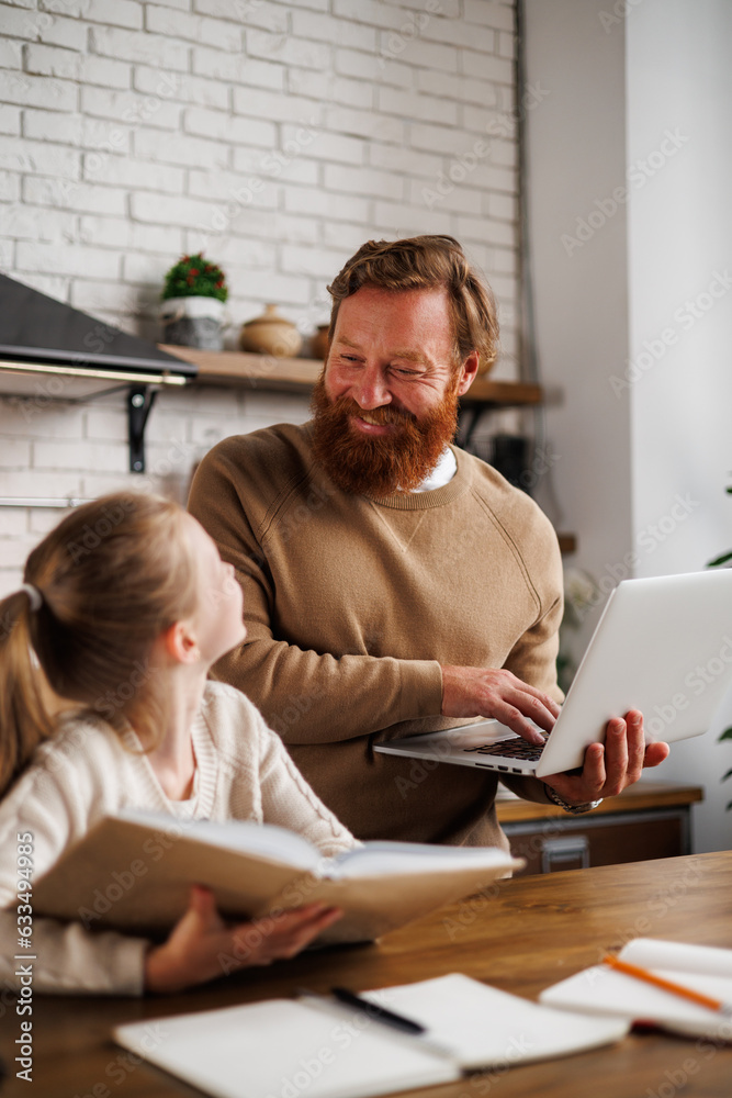 Positive dad holding laptop near daughter with book during education in kitchen at home Positive dad holding laptop near daughter with book during education in kitchen at home