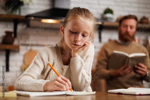 Freckled preadolescent girl writing on notebook near blurred father with book in kitchen at home