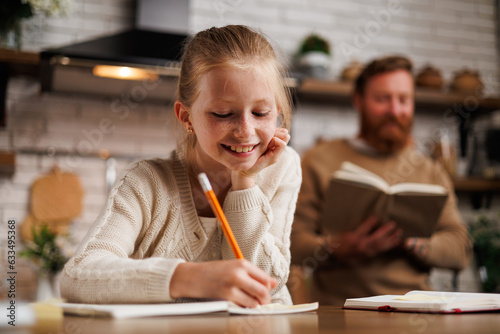 Smiling preadolescent kid writing on notebook near blurred father with book during homeschooling in kitchen  photo