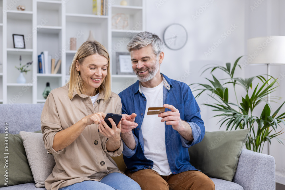Mature couple man and woman sitting together on sofa in living room at home, joyful family online choosing shopping products, using phone for online shopping and bank credit card Mature couple man and woman sitting together on sofa in living room at home, joyful family online choosing shopping products, using phone for online shopping and bank credit card