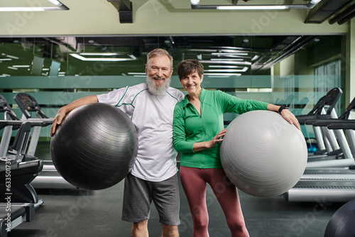 sporty elderly couple, cheerful man and woman holding fitness balls, active seniors in gym