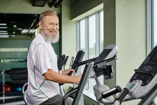 happy bearded man working out on exercise machine, elderly in gym, active senior, fitness and sport