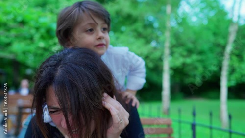 Abrasive little boy climbing on top of mother's neck harshly, showing little concern to mom's emotion in open air outside. parent in discomfort, parenting concept photo