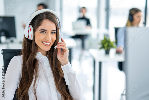Portrait of smiling long haired woman in headset working on computer and looking at camera in office.