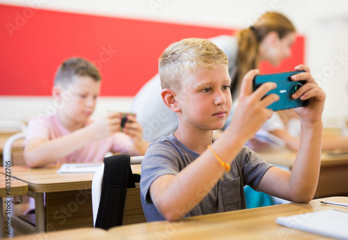 Schoolchildren sitting at desks in classroom. They're using their smartphones during lesson.