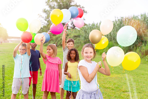 Portrait of happy preteen girl holding colorful helium balloons in hands, having fun with friends on green lawn on summer day